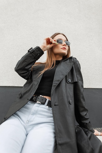 Mooi stijlvol model meisje in modieuze stedelijke vrijetijdskleding met een zwarte jas zwarte T-shirt en jeans zit en past haar zonnebril aan in de stad in de buurt van de muur