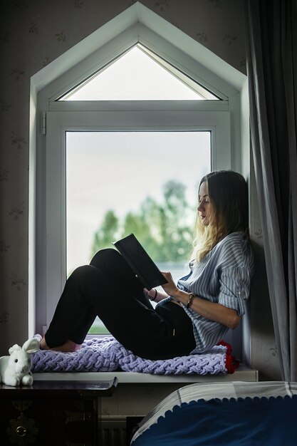 Mooi studentenmeisje zit thuis bij het raam en leest een boek