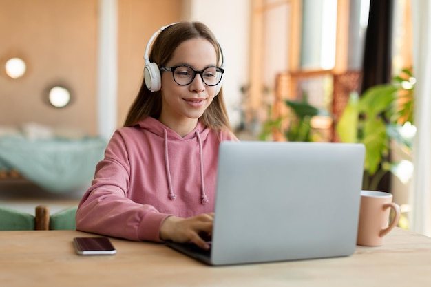 Mooi tienermeisje in draadloze headset met behulp van laptop geïnteresseerde gelukkige student typen op toetsenbord kijkend naar pc-scherm