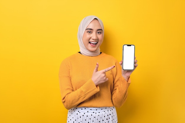 Mooi vrolijk Aziatisch meisje met hijab wijzende wijsvinger op mobiele telefoon met leeg wit scherm geïsoleerd op gele achtergrond mockup kopie ruimte