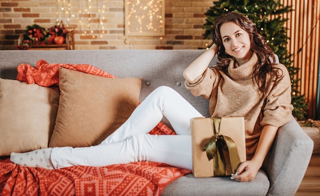 Mooi vrolijk gelukkig jong meisje met kerstcadeaus op de bank op de achtergrond van een nieuwjaarsboom thuis