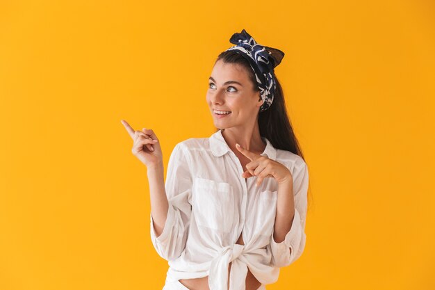 Mooi vrolijk jong meisje met zomerkleren die geïsoleerd over de gele muur staan en met de vingers naar de kopieerruimte wijzen