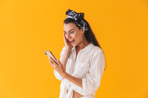 Mooi vrolijk jong meisje met zomerkleren die geïsoleerd over een gele muur staan, vieren tijdens het gebruik van mobiele telefoon