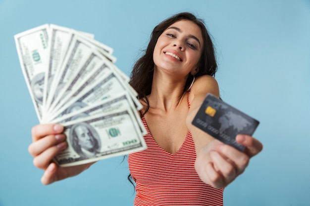 Mooi vrolijk meisje dat de zomerkleren draagt die zich geïsoleerd over blauw bevinden, geldbankbiljetten en creditcard tonen