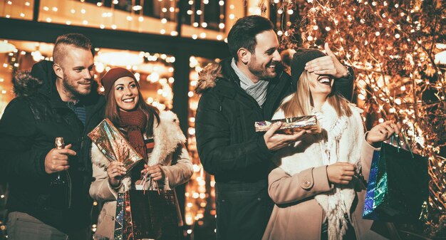 Mooi vrolijk stel dat Kerstmis viert in de stadsstraat. Jonge man die de ogen van een blij verraste vriendin bedekt en haar cadeau geeft.