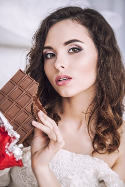 Mooi vrouwenmodel met tegels van donkere chocolade in handen tegen slaapkamerbinnenland