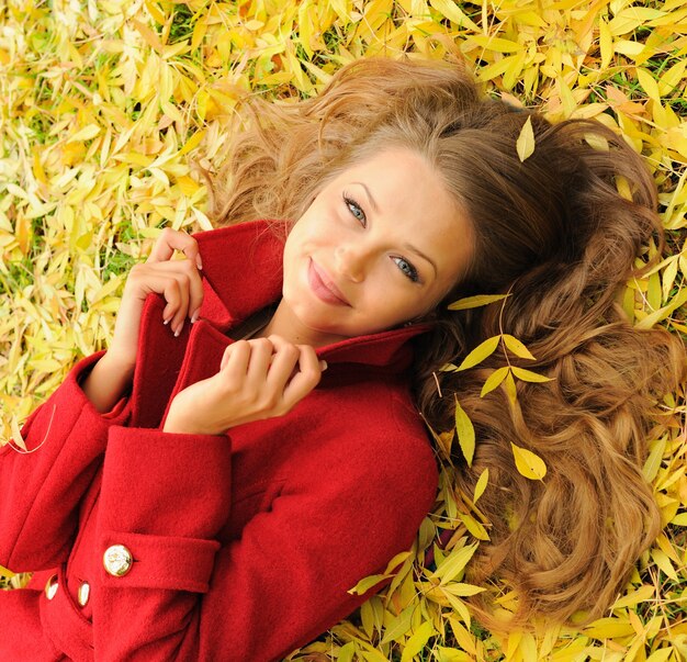 Mooi vrouwenportret dat op gele esdoornbladeren in park ligt, gekleed in mode rode jas