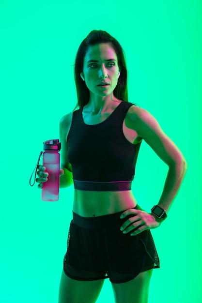 Mooi zelfverzekerd jong fitnessmeisje dat geïsoleerd staat over een groene neonmuur, draadloze oortelefoons draagt en een waterfles vasthoudt na de training