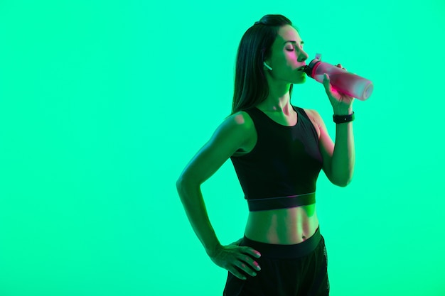 Mooi zelfverzekerd jong fitnessmeisje dat geïsoleerd staat over groene neonmuur, draadloze oortelefoons draagt, water drinkt uit een fles na de training