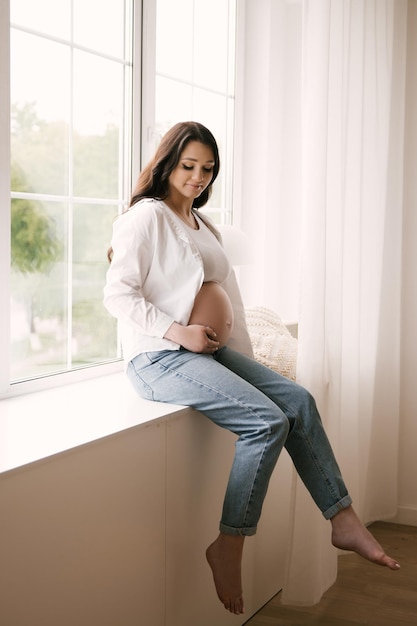Mooi zwanger meisje met krullen in een wit overhemd en jeans in een lichte studio met een stijlvol interieur het concept van een gelukkige zwangerschap en familie