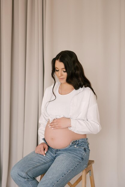 Mooi zwanger meisje met krullen in een wit overhemd en jeans in een lichte studio met een stijlvol interieur het concept van een gelukkige zwangerschap en familie