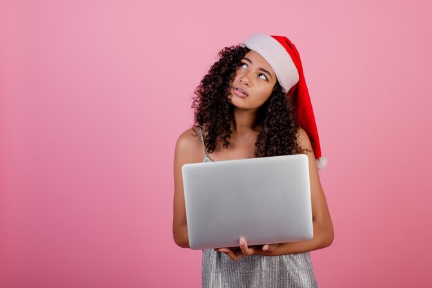 Mooi zwart meisje met laptop die de hoed en de kleding van vakantiekerstmis draagt die over roze wordt geïsoleerd