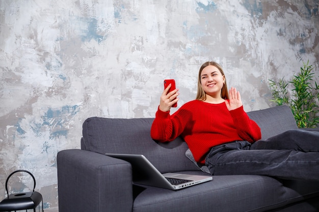 Mooie aantrekkelijke jonge vrouw zit thuis op de bank en werkt aan de telefoon, ze is gekleed in een rode trui en jeans, naast een laptop
