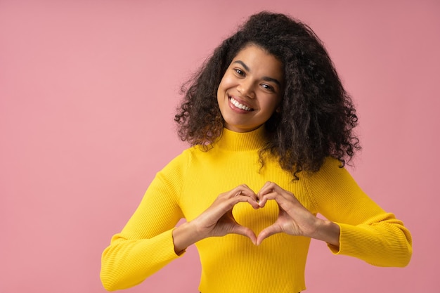 Mooie Afrikaanse Amerikaanse vrouw met brede glimlach die hartvorm toont die op roze achtergrond wordt geïsoleerd