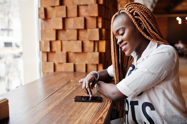 Mooie Afrikaanse vrouw in stijlvolle casual shirt en dreadlocks poseren in café in de buurt van de vensterbank en haar mobiele telefoon aan te raken.