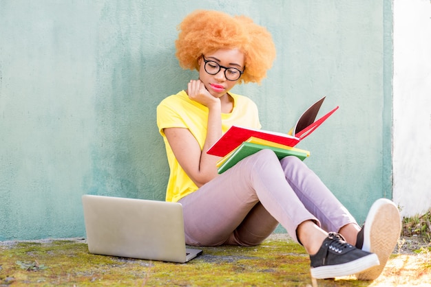 Mooie Afrikaanse vrouw met krullend haar studeren met boeken en laptop buiten zitten op de groene muur achtergrond