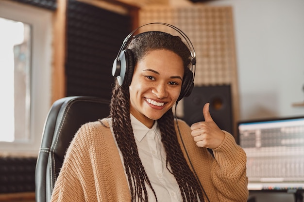 Mooie afrikaanse vrouw muzikant in koptelefoon toont duim omhoog glimlacht brede glimlach kijkend naar camera