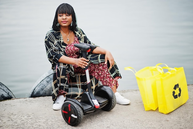 Mooie afro-amerikaanse vrouw zit in de buurt van segway of hoverboard Zwart meisje met gele stoffen eco-tassen recyclingsymbool