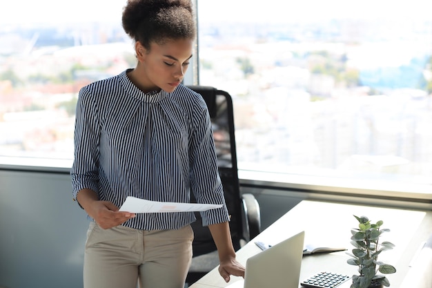 Mooie afro-amerikaanse zakenvrouw in slimme vrijetijdskleding die op laptop op kantoor werkt.