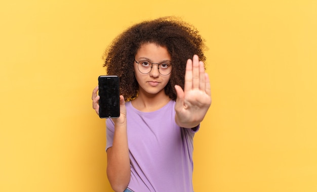 Mooie afro-tiener die er serieus, streng, ontevreden en boos uitziet met een open palm die een stopgebaar maakt en een cel vasthoudt