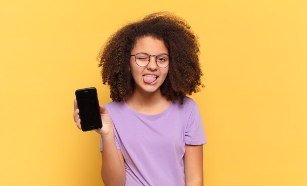 Mooie afro-tiener met vrolijke, zorgeloze, rebelse houding, grappen maken en tong uitsteken, plezier maken en een cel vasthouden