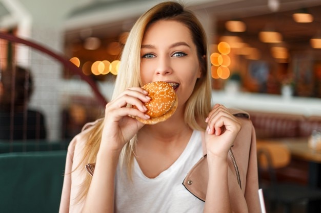 Mooie Amerikaanse jonge vrouw in een roze jasje met een hamburger in een restaurant