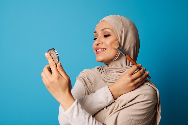 Mooie Arabische moslimvrouw in hijab houdt een cosmetische spiegel en een make-upborstel en geldt blush op de jukbeenderen van haar gezicht, geïsoleerd