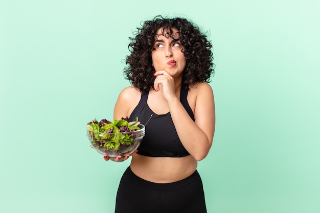 Mooie arabische vrouw die denkt, zich twijfelachtig en verward voelt en een salade vasthoudt. dieet concept