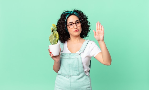 Mooie arabische vrouw die er serieus uitziet en een open palm toont die een stopgebaar maakt en een ingemaakte cactus vasthoudt