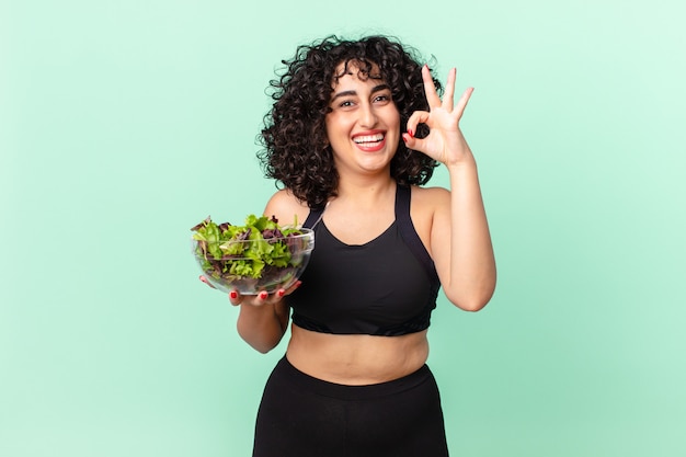 Mooie arabische vrouw die zich gelukkig voelt, goedkeuring toont met een goed gebaar en een salade vasthoudt. dieet concept