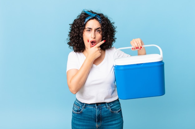 Mooie Arabische vrouw met wijd open mond en ogen en hand op kin en met een draagbare koelkast