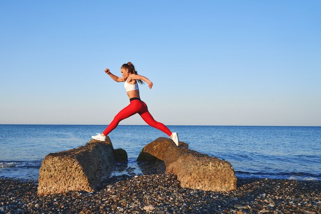 Mooie atleet vrouw in rode sportkleding poseren op de achtergrond van het landschap van de zee. Ochtendtraining bij de oceaan, gezonde levensstijl