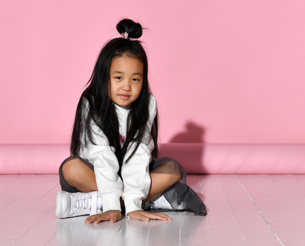 Mooie Aziatische brunette meisje zittend in studio op roze achtergrond en kijken naar camera Kind met een mooie verschijning gekleed in stijlvolle kleding Concept van kindermode en etniciteit