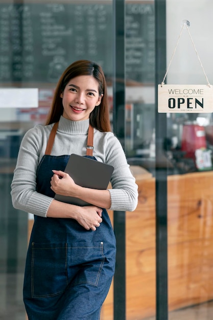 Mooie aziatische jonge barista-vrouw in schort die tablet vasthoudt en voor de deur van café staat met open bord Bedrijfseigenaar opstarten MKB-ondernemerconcept