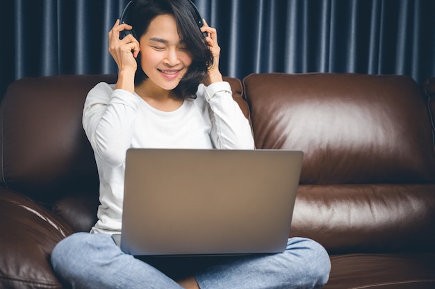 Foto mooie aziatische jonge vrouw ontspannen met haar muziek spelen met laptop op de bank. thuis werken en geluk concept.