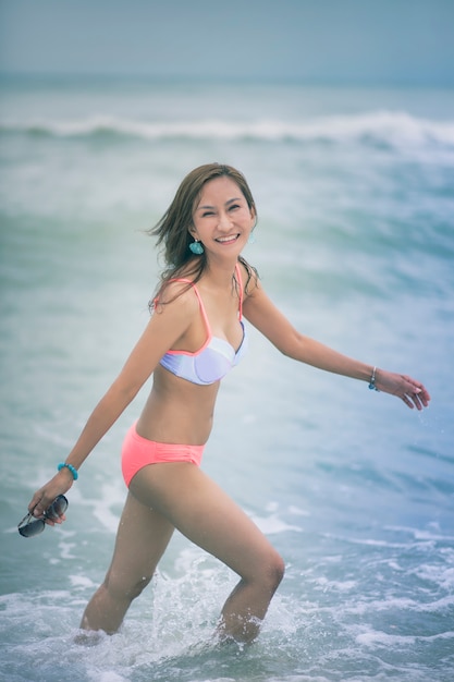 Mooie Aziatische jongere vrouw die bikini op overzees strand met het glimlachen gezicht draagt