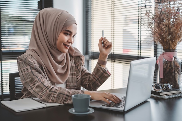 Mooie Aziatische moslim zakelijke vrouw bruine hijab zitten en werken met laptopcomputer op moderne kantoor.