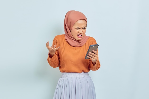 Mooie Aziatische moslimvrouw in bruine trui en hijab met mobiele telefoon met boze uitdrukking geïsoleerd op een witte achtergrond moslim lifestyle concept