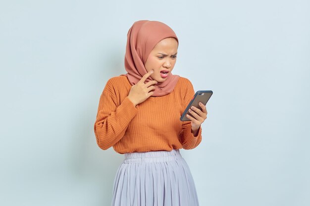 Mooie Aziatische Moslimvrouw in bruine trui en hijab met mobiele telefoon met verwarde uitdrukking geïsoleerd op een witte achtergrond Moslim levensstijl concept