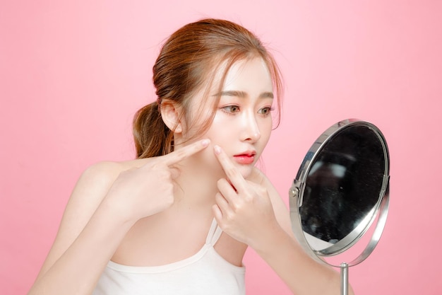Mooie Aziatische schoonheid vrouw model Pony haar met kijk in de spiegel en zorgen over acne rimpels op het gezicht en perfecte huid op geïsoleerde roze achtergrond gezichtsbehandeling cosmetologie plastische chirurgie
