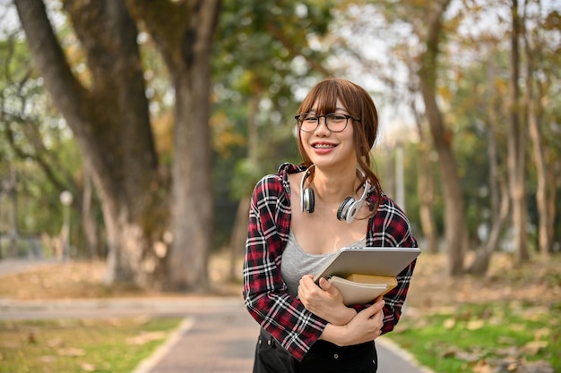 Mooie Aziatische studente die haar laptop en schoolboek vasthoudt terwijl ze langs het park loopt