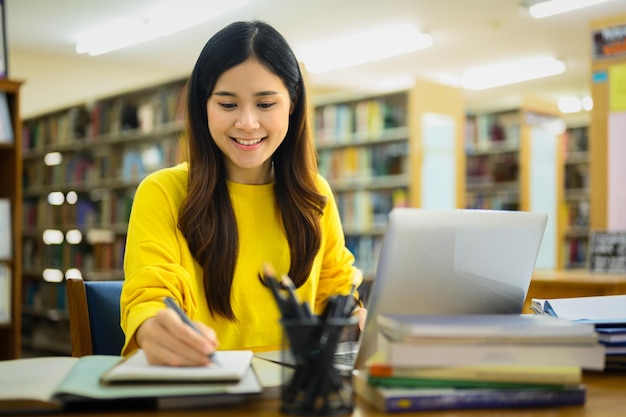 Mooie Aziatische universiteitsvrouw die rapport voorbereiden dat opdrachten op laptop in een bibliotheek doet