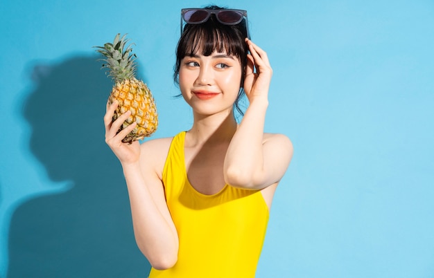 Mooie Aziatische vrouw die gele jumpsuit op blauw draagt en tropisch fruit eet