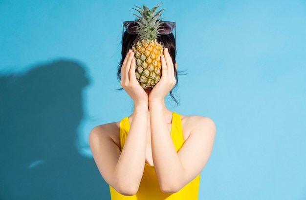 Mooie Aziatische vrouw die gele jumpsuit op blauw draagt en tropisch fruit eet