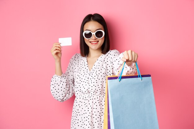 Mooie aziatische vrouw die in zonnebril gaat winkelen, zakken vasthoudt en creditcard toont, die zich over roze bevindt