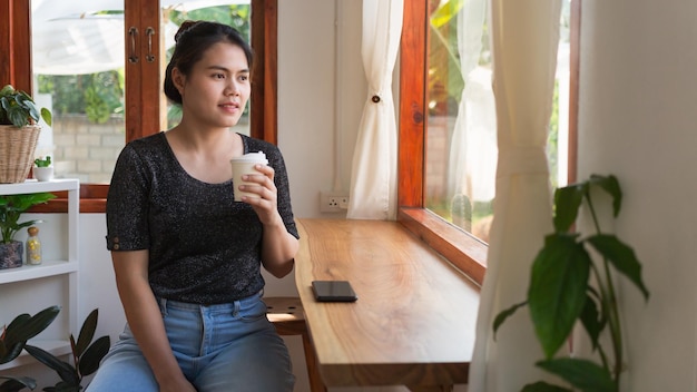 Mooie Aziatische vrouw gaat aan de bar zitten met een papieren koffiekopje glimlachend ontspannen in café