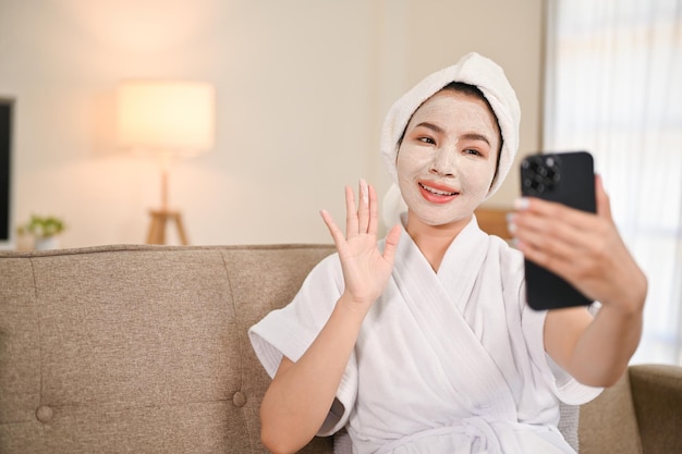 Mooie Aziatische vrouw in badjas videogesprek met vrienden terwijl ze thuis een gezichtsmasker aanbrengt