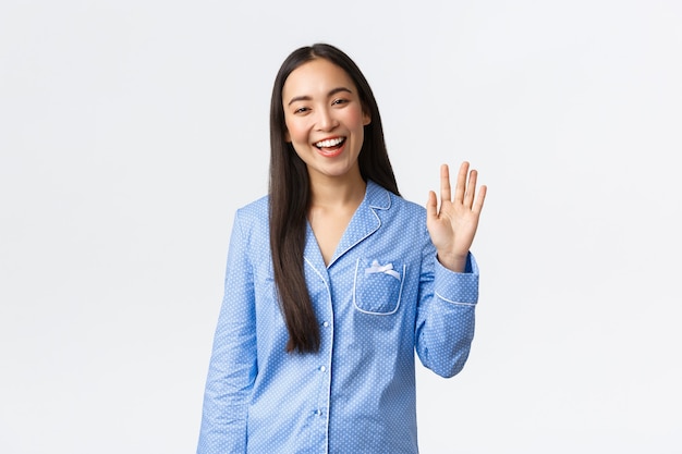Mooie Aziatische vrouw in blauwe pyjama glimlachend gelukkig en zwaaiende hand om hallo te zeggen, groet vriendin en welkom bij sleepover party, staande witte achtergrond met hallo gebaar, witte achtergrond.