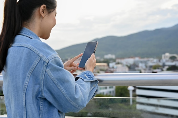 Mooie Aziatische vrouw in spijkerjasje met behulp van haar smartphone op het dak van het achteraanzicht van het gebouw