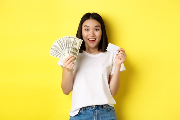 Mooie aziatische vrouw met kort donker haar, gekleed in wit t-shirt, met geld in dollars en plastic creditcard, staande over gele achtergrond